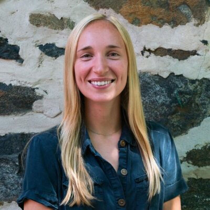 The image shows a woman with long blonde hair, smiling at the camera. She is wearing a dark blue button-down shirt. The background is a stone wall with various shades of gray and brown. The lighting appears to be natural, suggesting the photo was taken outdoors. The woman's expression is friendly and approachable.